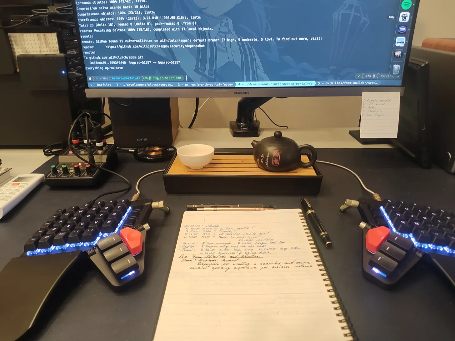 Split keyboard with a notebook in the middle, a teapot, a small cup and half of a monitor showing code on it